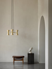 A brown leather Elizabeth Ottoman and a black marble Plinth Tall sit under a Collector Chandelier in a minimalist room.