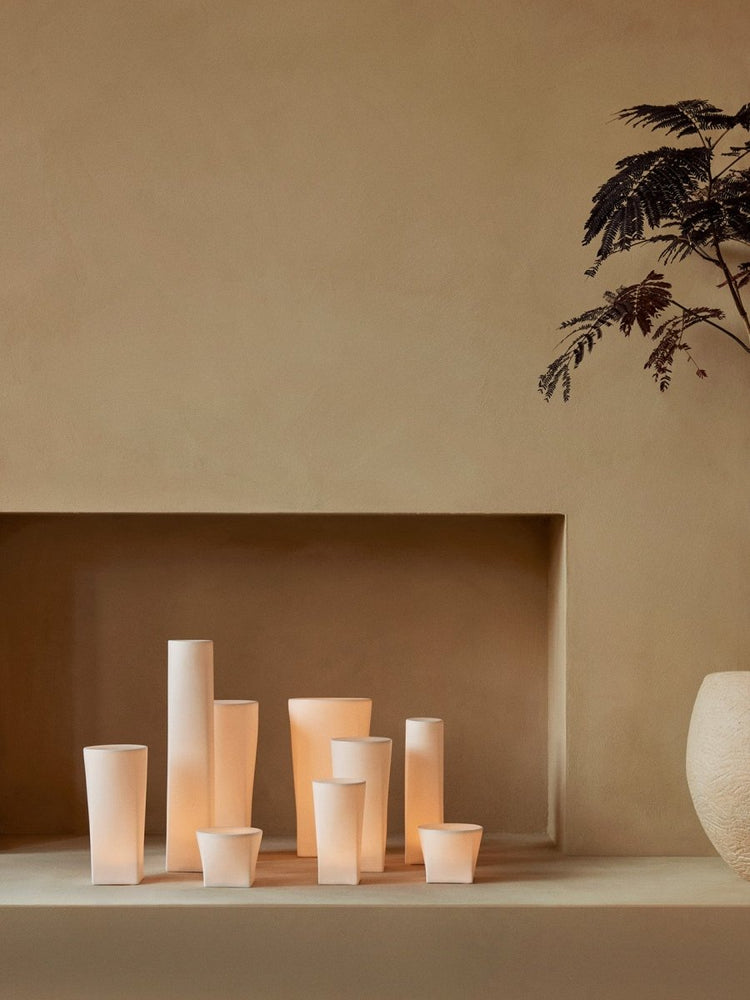 Various sizes of Ignus Flameless Candles glow warmly on a minimalist beige shelf next to a Plantas Planter and dark plant.
