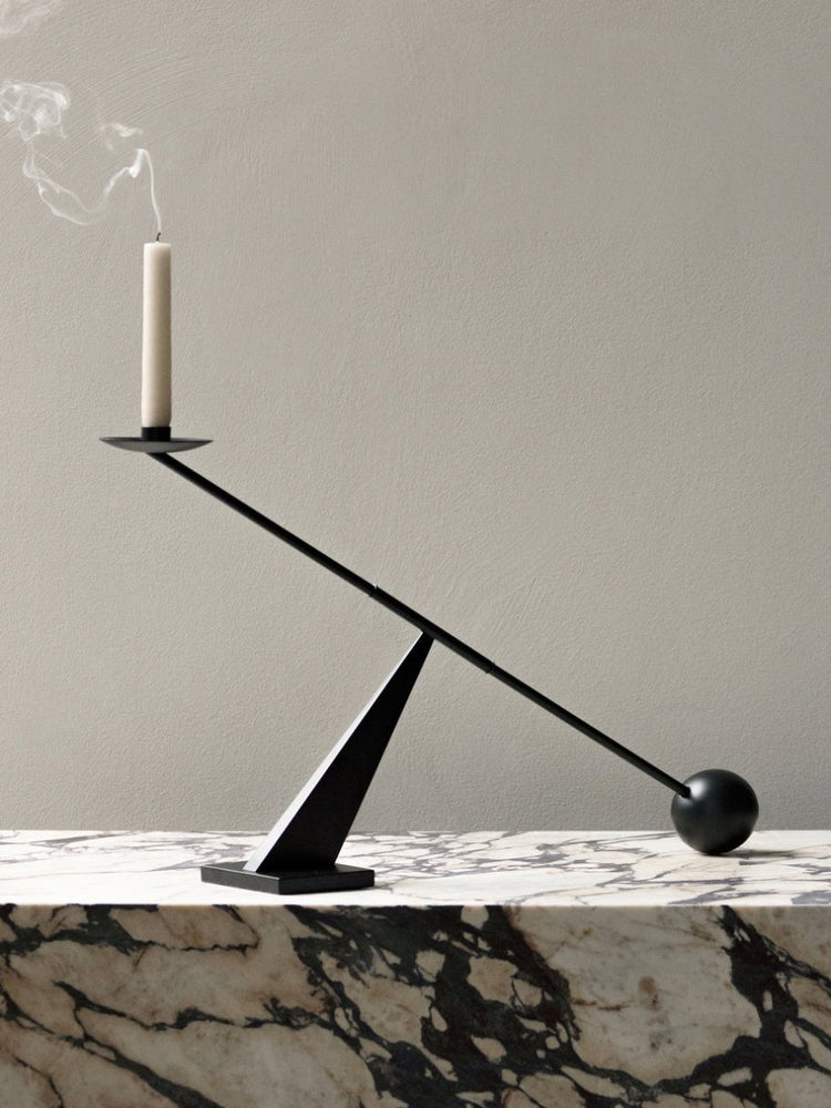 A black Interconnect Candleholder balancing an extinguished white candle on a marble Plinth against a grey wall.