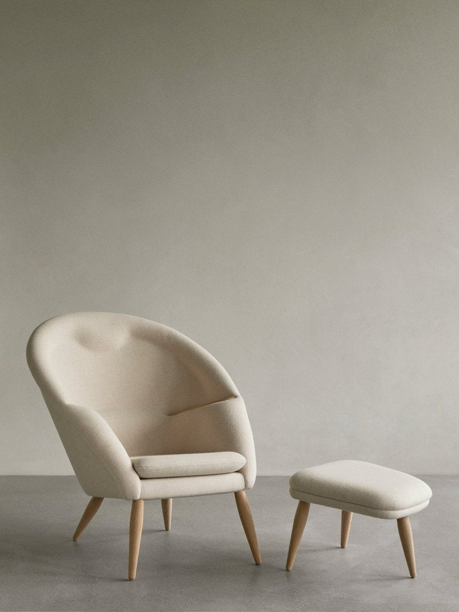 A cream-colored Oda Lounge Chair with a curved back and oak legs sits next to the matching Oda Ottoman on a gray floor.