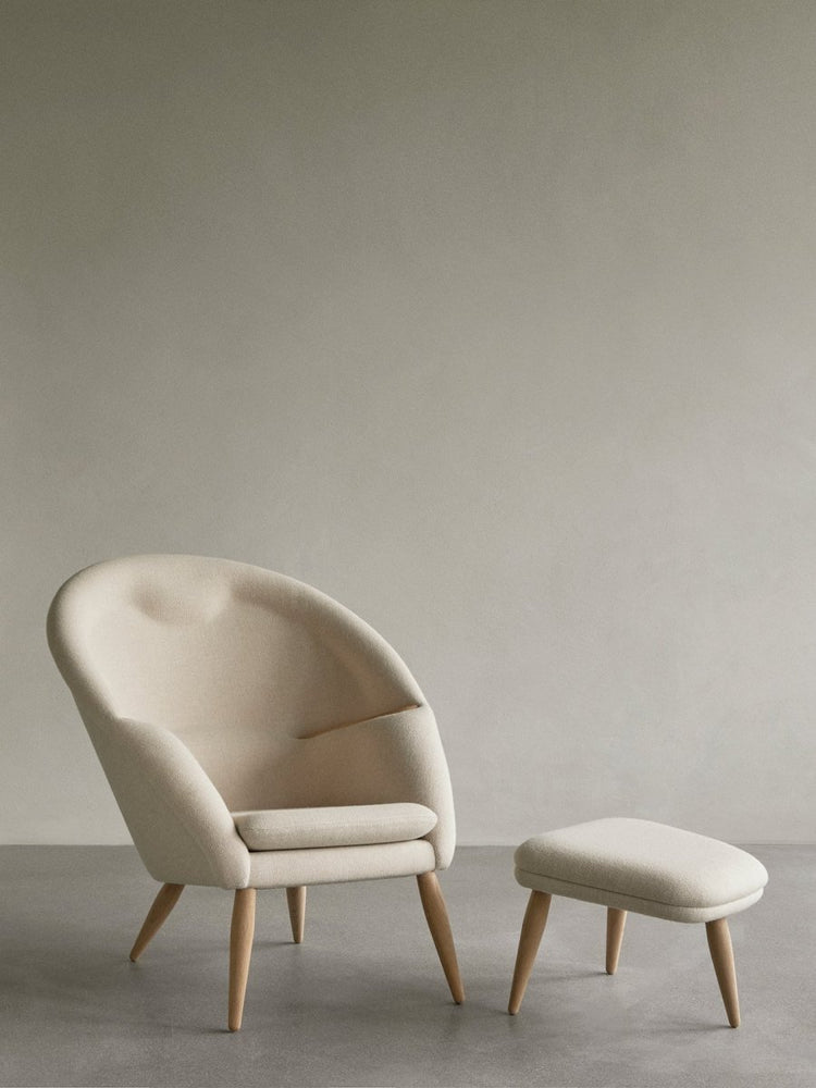 A cream-colored Oda Lounge Chair with a curved back and oak legs sits next to the matching Oda Ottoman on a gray floor.