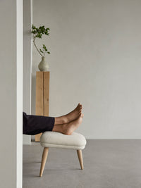 A person's bare feet rest on the Oda Ottoman with light fabric and natural oak legs in a minimal room with a wooden pedestal.