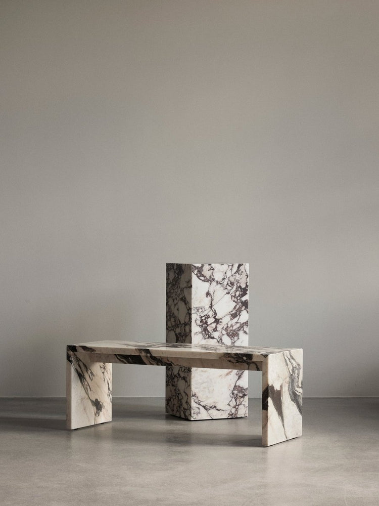 The Plinth Bridge and Plinth Pedestal in heavily veined marble, staged in a minimalist room with a gray wall and floor.