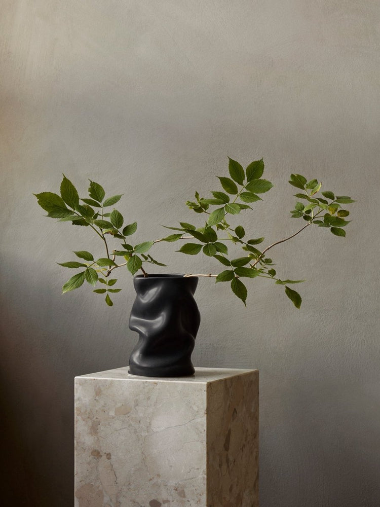 Sculptural, matte black Collapse Vase holds leafy green branches, displayed on a marble Plinth Pedestal against a gray wall.