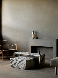 The Seal Lounge Chair next to veined marble Plinth Bridge and Plinth Low tables in a modern gray-toned room by a fireplace.