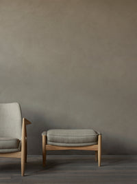 The Seal Ottoman in gray fabric and oak sits next to the Seal Lounge Chair on dark wood floors against a textured taupe wall.