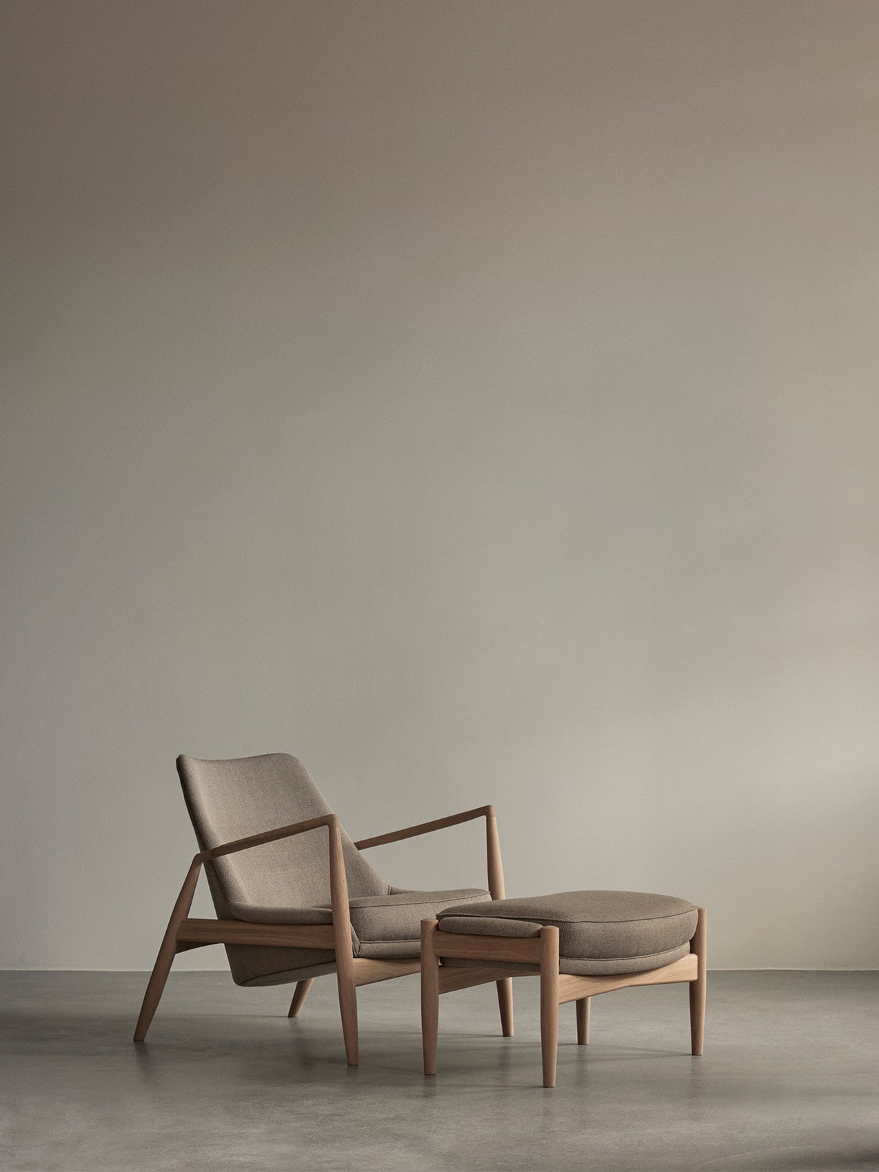 Beige upholstered lounge chair with oak frame and matching ottoman on concrete floor against a neutral taupe wall