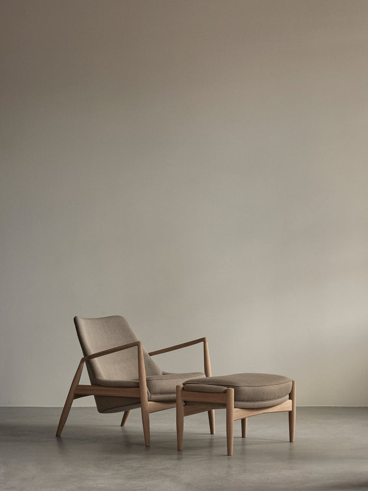 Beige upholstered lounge chair with oak frame and matching ottoman on concrete floor against a neutral taupe wall