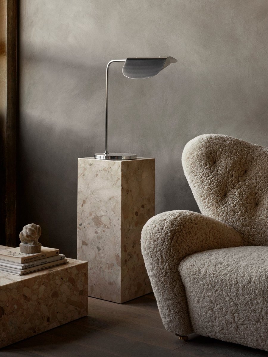 Chrome Wing Table Lamp on a marble Plinth Pedestal next to a cream shearling Tired Man armchair against a textured gray wall.