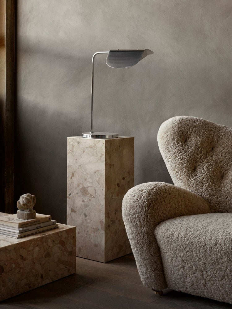 Chrome Wing Table Lamp on a marble Plinth Pedestal next to a cream shearling Tired Man armchair against a textured gray wall.