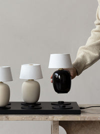 A hand in a beige sweater placing a black Torso Portable Table Lamp onto a Charging Board on a marble Androgyne Lounge Table.