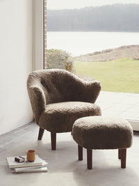 Ingeborg Lounge Chair and matching Ottoman in brown Sahara sheepskin, staged in a sunlit room overlooking a tranquil lake.
